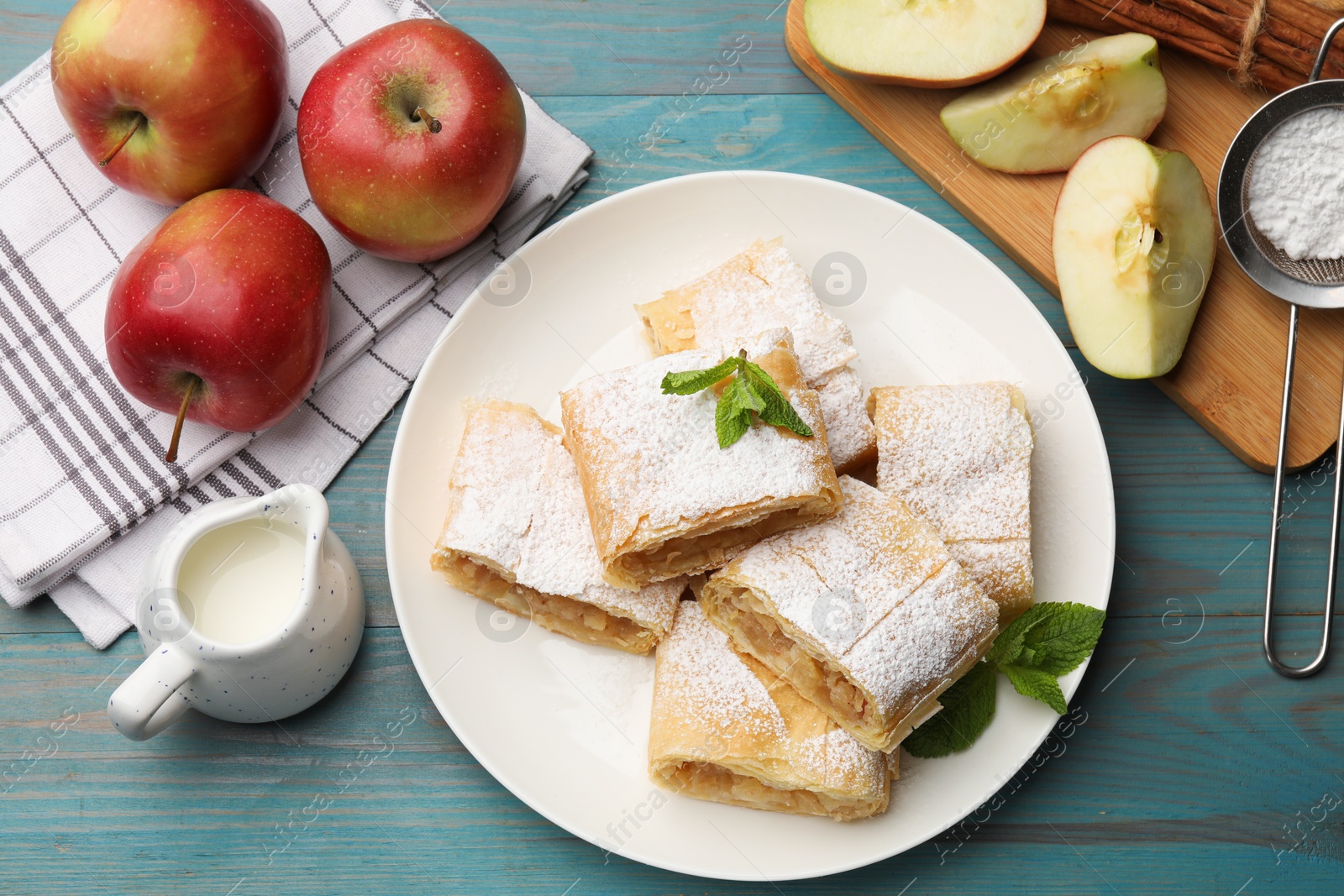 Pieces of tasty apple strudel with powdered sugar, mint and fruits on light blue wooden table, flat lay Photo of Pieces of tasty apple strudel with powdered sugar, mint and fruits on light blue wooden table, flat lay