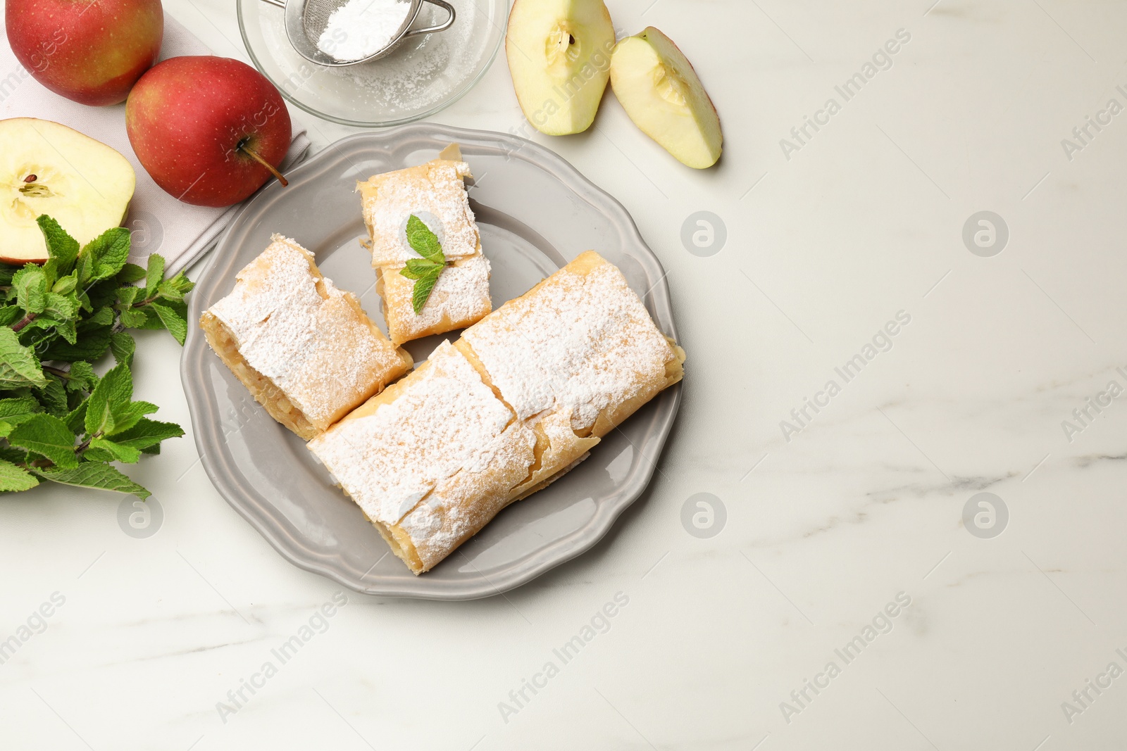 Photo of Pieces of tasty apple strudel with powdered sugar, mint and fruits on white marble table, flat lay
