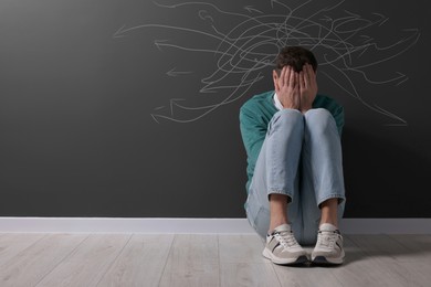Image of Upset man sitting on floor. Confused thoughts depicted by lines and arrows on black wall behind him. Stress, anxiety, depression