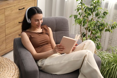 Photo of Harmony. Smiling woman in headphones reading book on armchair at home