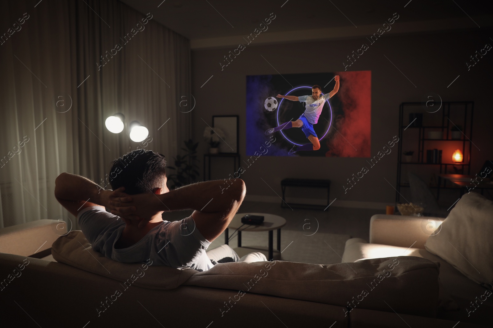 Man watching sports event using video projector on sofa at night Image of Man watching sports event using video projector on sofa at night