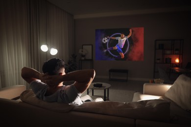 Image of Man watching sports event using video projector on sofa at night