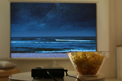 Image of Video projector and bowl with chips on wooden table. Night seascape on projection screen