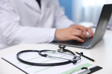 Photo of Doctor using laptop at table in clinic, focus on clipboard and stethoscope. Online medical consultation
