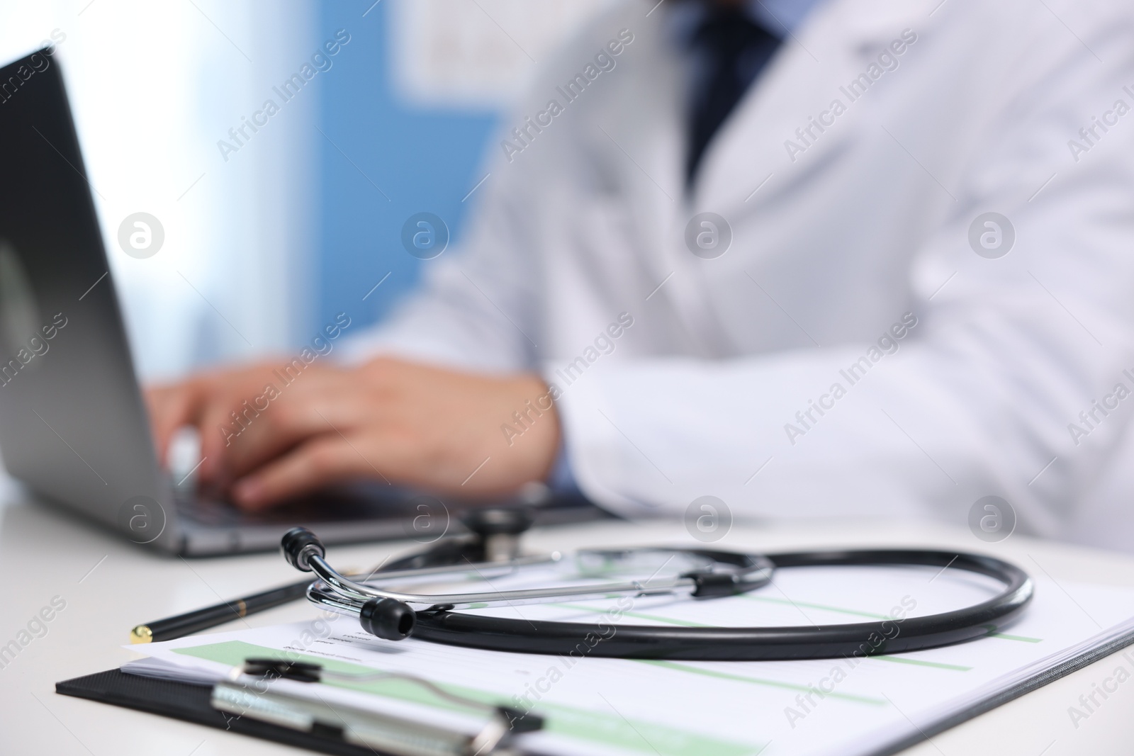 Doctor using laptop at table in clinic, focus on clipboard and stethoscope. Online medical consultation Photo of Doctor using laptop at table in clinic, focus on clipboard and stethoscope. Online medical consultation