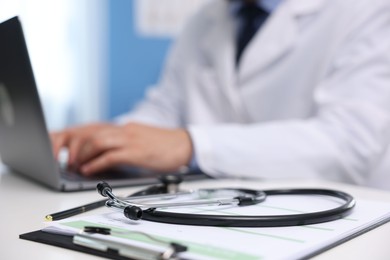 Photo of Doctor using laptop at table in clinic, focus on clipboard and stethoscope. Online medical consultation