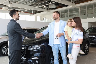 Photo of Salesman shaking hands with happy family near new car in salon