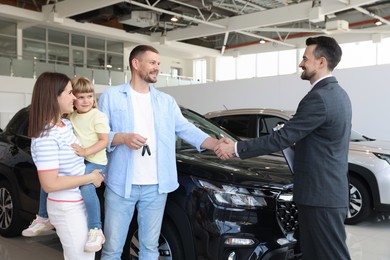 Photo of Salesman shaking hands with happy family near new car in salon