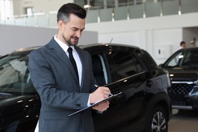 Photo of Smiling salesman working with clipboard near new car in salon