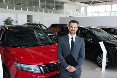 Photo of Portrait of smiling salesman with clipboard near new car in salon