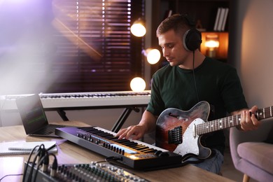Photo of Man in headphones working with MIDI keyboard and guitar at table in home studio
