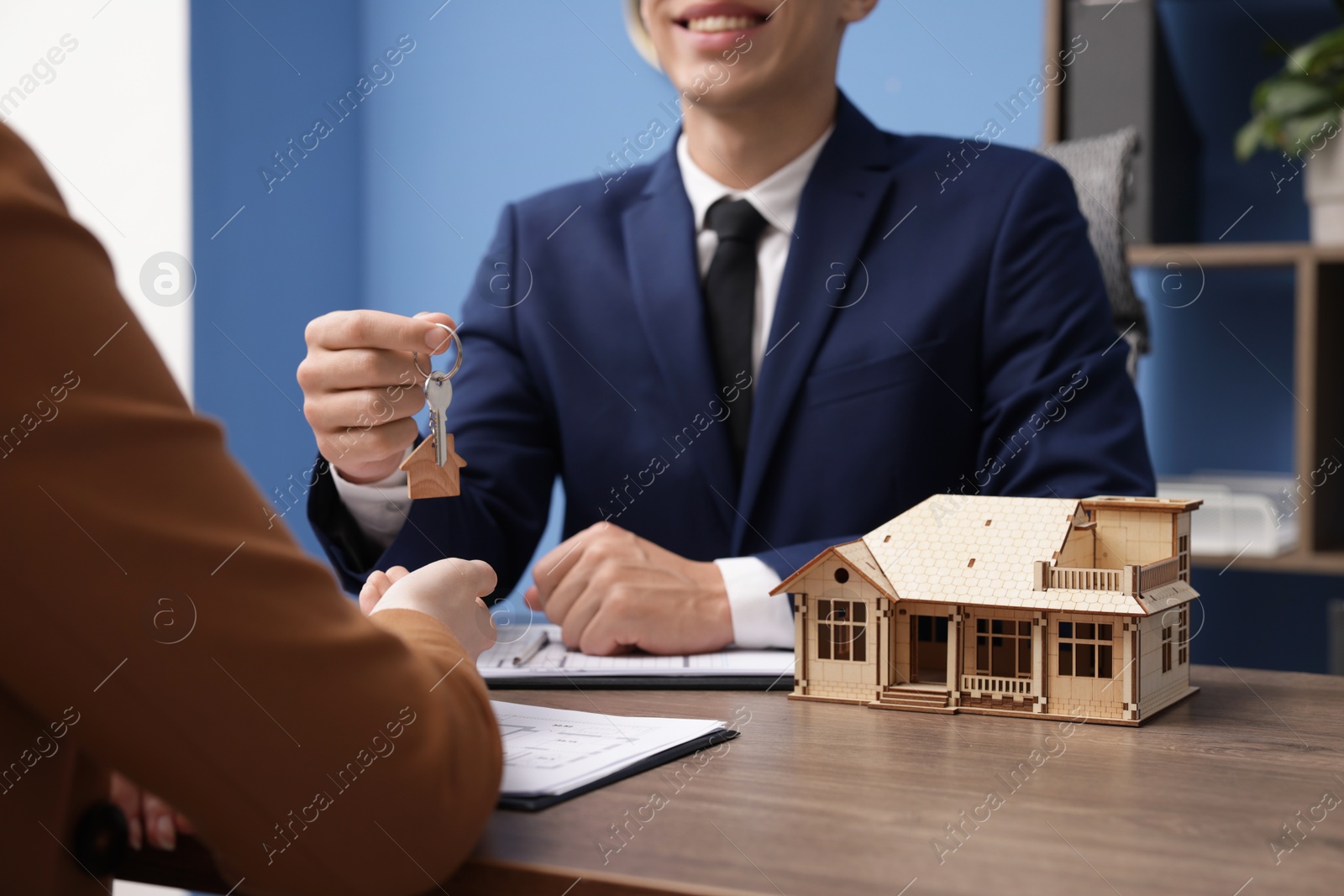 Real estate agent giving key to client at desk in office, focus on house model Photo of Real estate agent giving key to client at desk in office, focus on house model