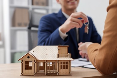 Photo of Real estate agent giving key to client at desk in office, focus on house model