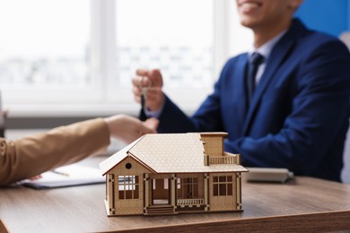 Photo of Real estate agent giving key to client at desk in office, focus on house model