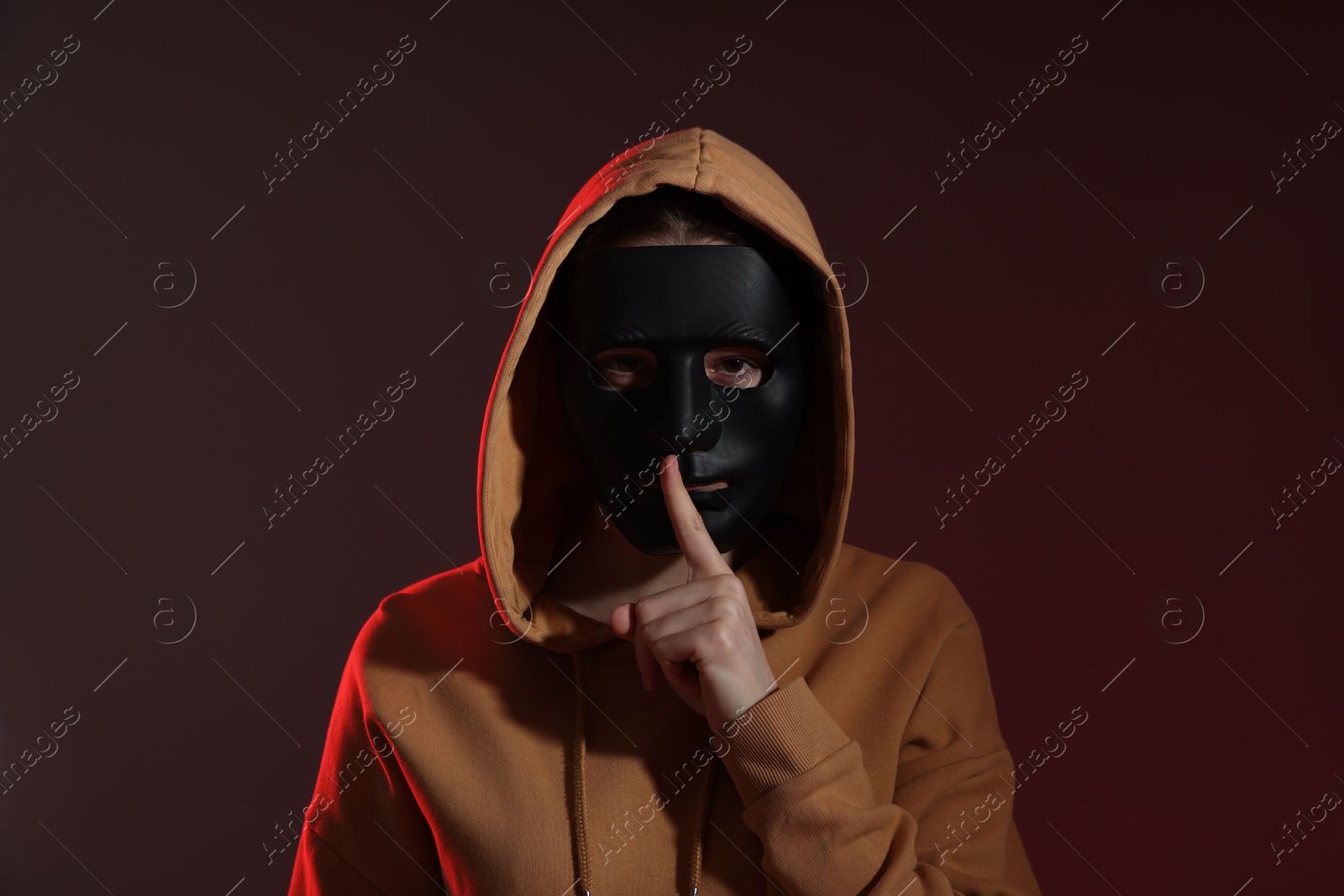 Photo of Anonymity. Woman wearing mask and showing hush gesture on dark background