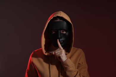 Photo of Anonymity. Woman wearing mask and showing hush gesture on dark background