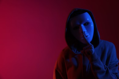 Photo of Anonymity. Woman wearing mask and showing hush gesture on burgundy background in blue light, space for text