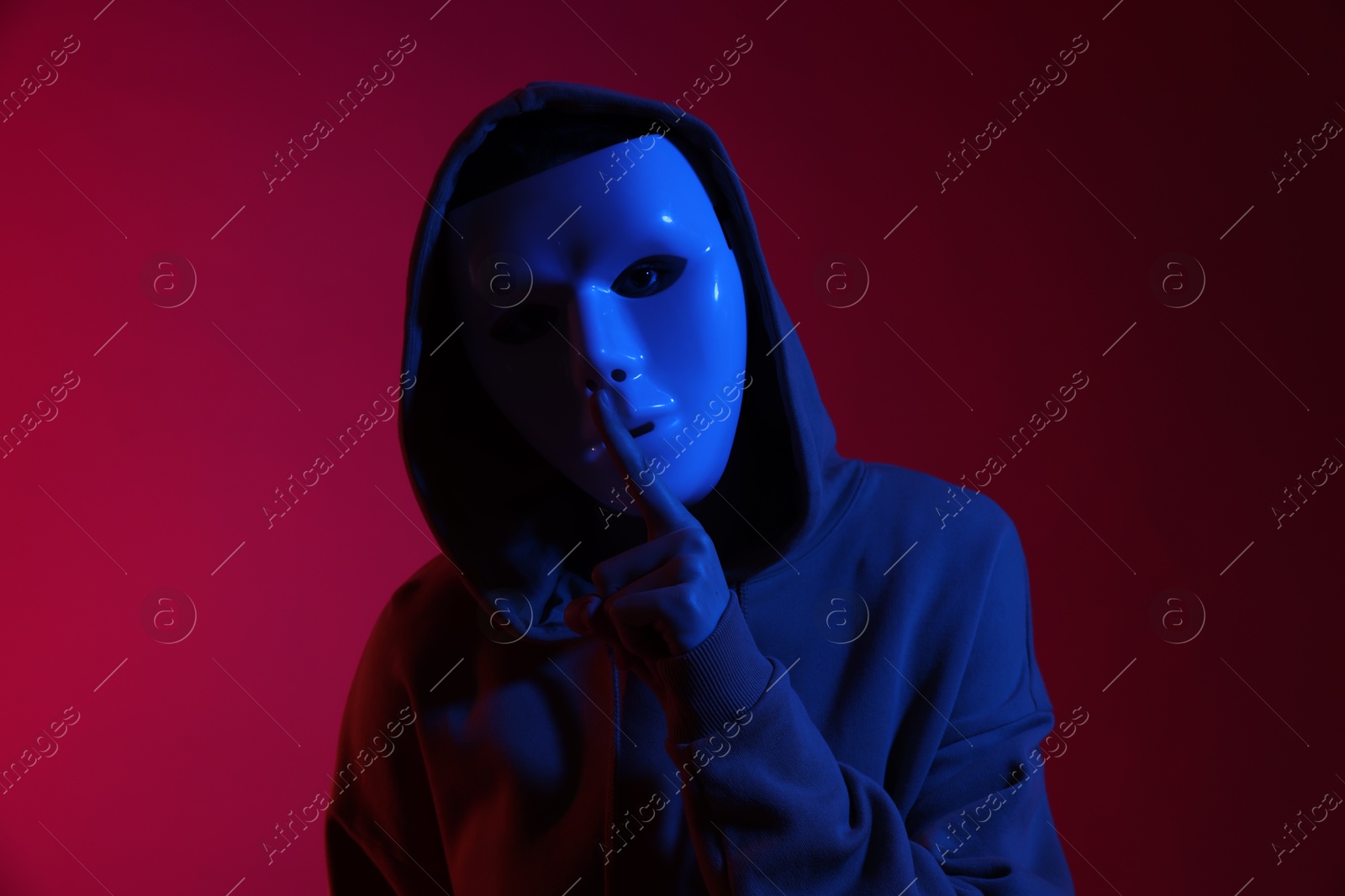Photo of Anonymity. Woman wearing mask and showing hush gesture on burgundy background in blue light