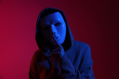 Photo of Anonymity. Woman wearing mask and showing hush gesture on burgundy background in blue light
