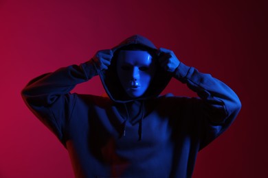 Photo of Anonymity. Woman wearing mask on burgundy background in blue light