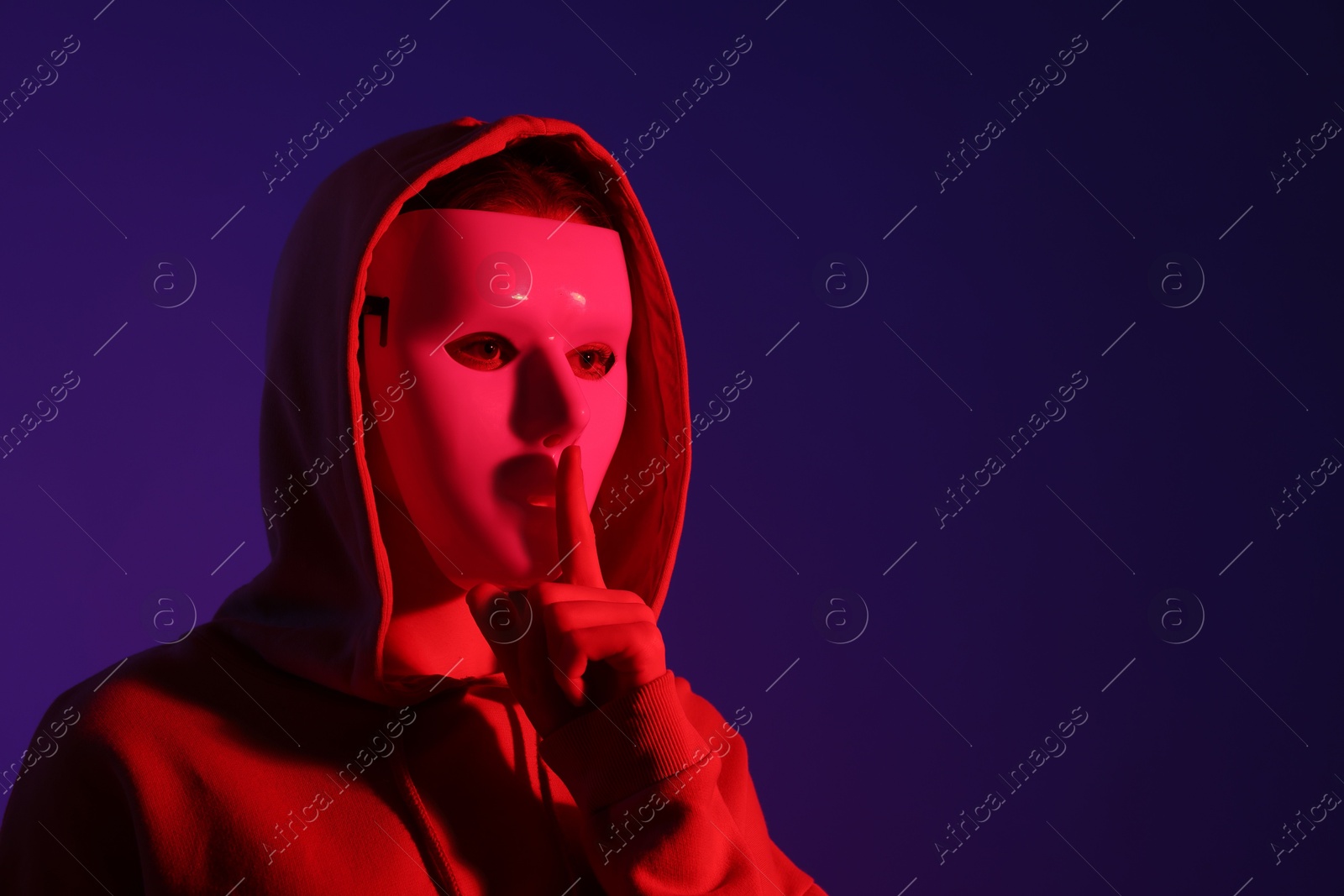 Photo of Anonymity. Woman wearing mask and showing hush gesture on dark blue background in red light, space for text