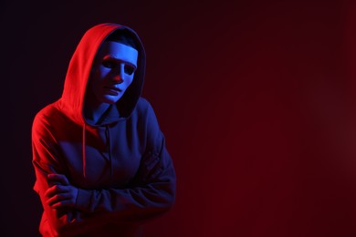 Photo of Anonymity. Woman wearing mask on dark red background in neon lights, space for text