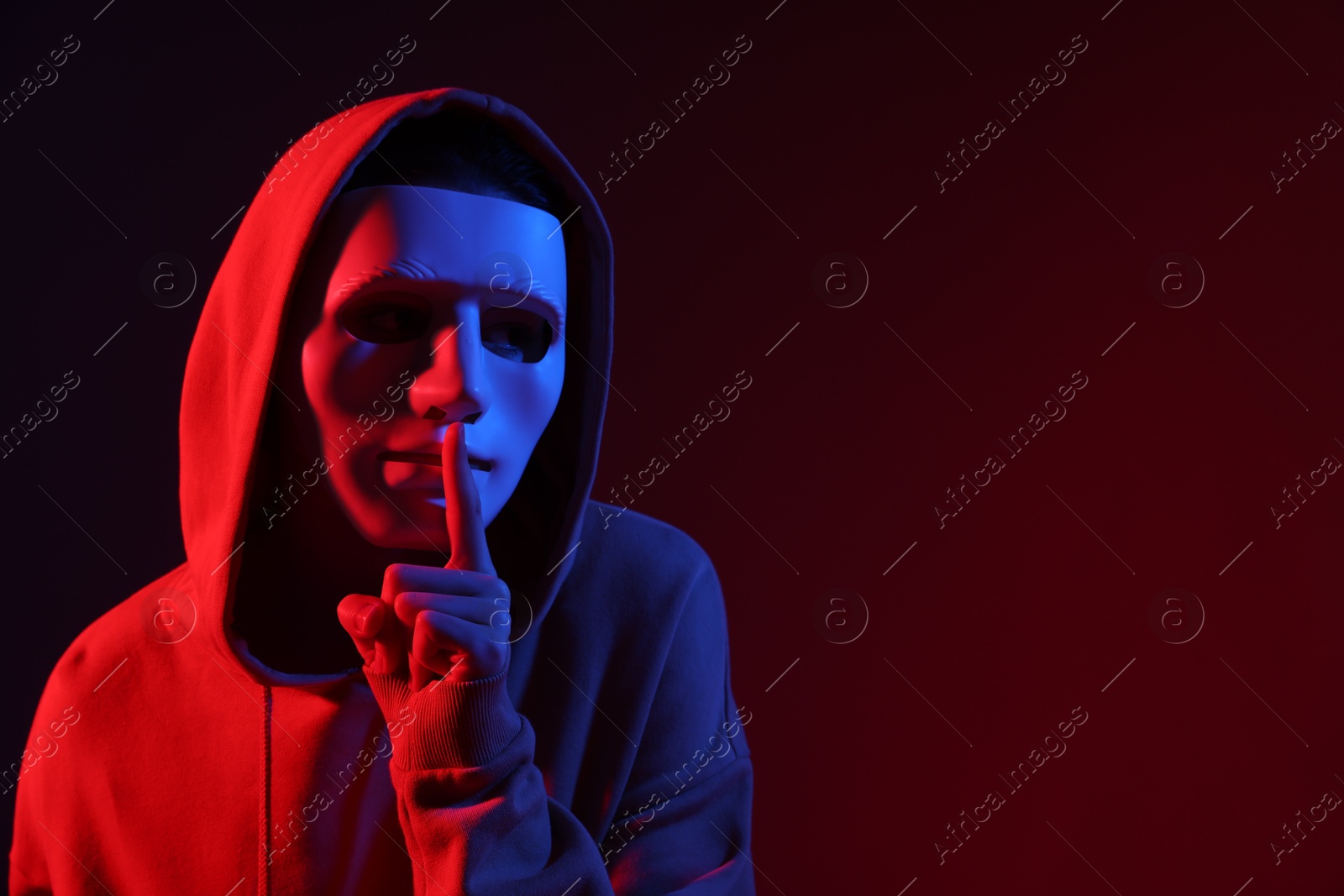 Photo of Anonymity. Woman wearing mask and showing hush gesture on dark red background in neon lights, space for text