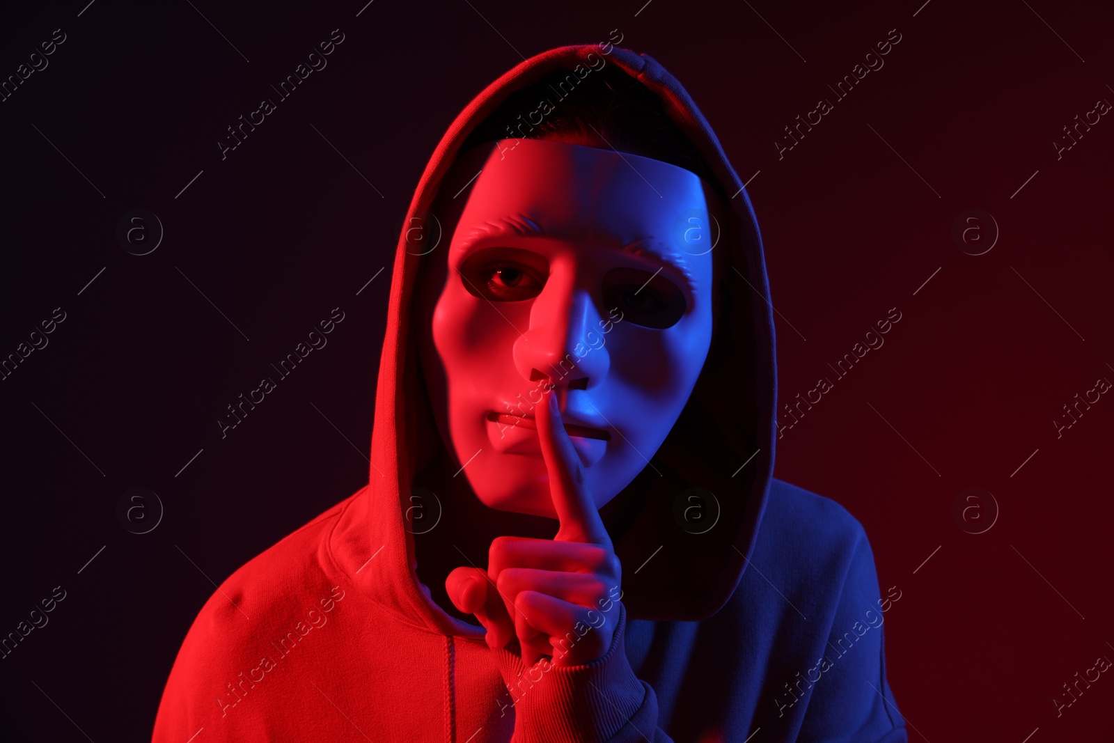 Photo of Anonymity. Woman wearing mask and showing hush gesture on dark red background in neon lights