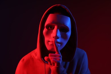 Photo of Anonymity. Woman wearing mask and showing hush gesture on dark red background in neon lights