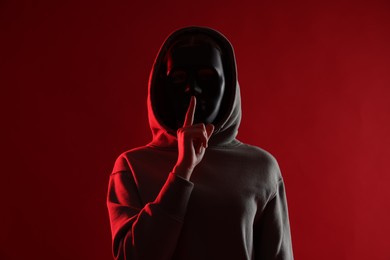 Photo of Anonymity. Woman wearing mask and showing hush gesture on red background
