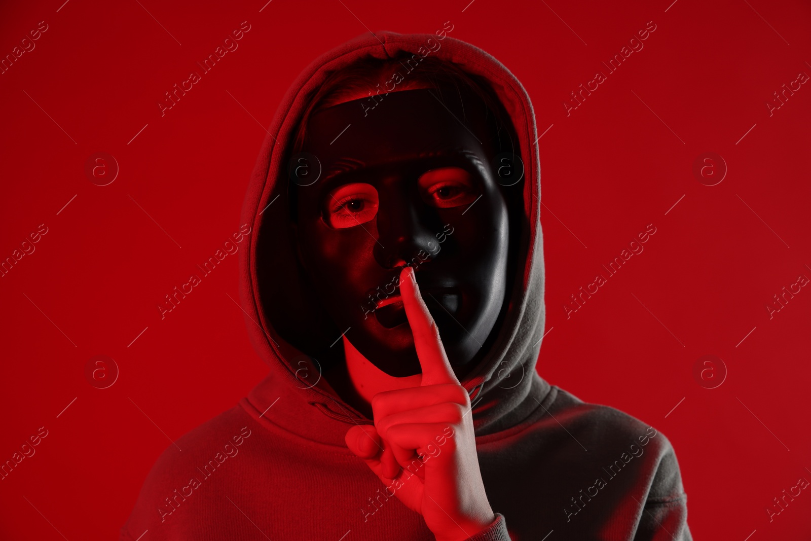 Photo of Anonymity. Woman wearing mask and showing hush gesture on red background
