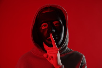 Photo of Anonymity. Woman wearing mask and showing hush gesture on red background