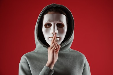 Photo of Anonymity. Woman wearing mask and showing hush gesture on red background