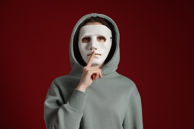 Photo of Anonymity. Woman wearing mask and showing hush gesture on dark red background