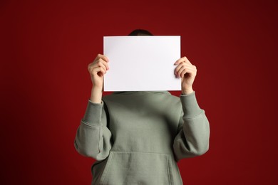 Photo of Anonymity. Woman hiding behind blank paper sheet on dark red background