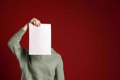 Photo of Anonymity. Woman hiding behind blank paper sheet on dark red background, space for text