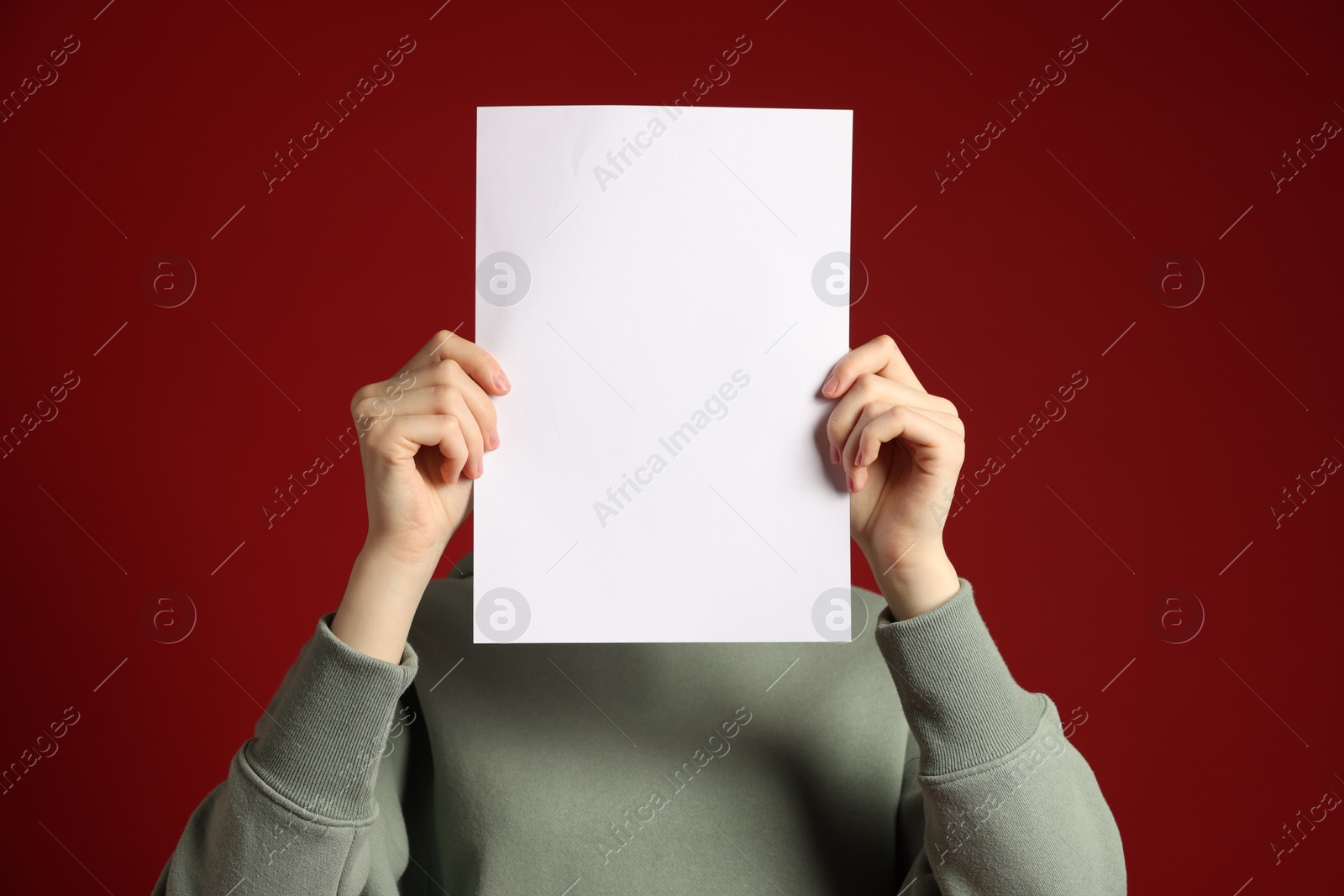 Anonymity. Woman hiding behind blank paper sheet on dark red background Photo of Anonymity. Woman hiding behind blank paper sheet on dark red background