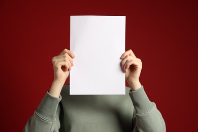 Photo of Anonymity. Woman hiding behind blank paper sheet on dark red background