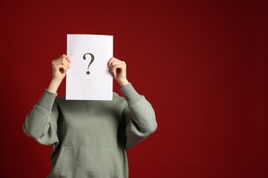 Photo of Anonymity. Woman hiding behind paper sheet with question mark on dark red background, space for text
