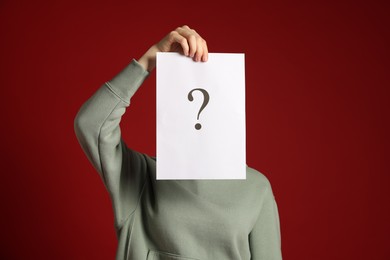 Photo of Anonymity. Woman hiding behind paper sheet with question mark on dark red background