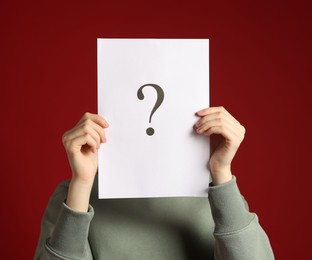 Photo of Anonymity. Woman hiding behind paper sheet with question mark on dark red background