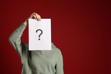 Photo of Anonymity. Woman hiding behind paper sheet with question mark on dark red background, space for text