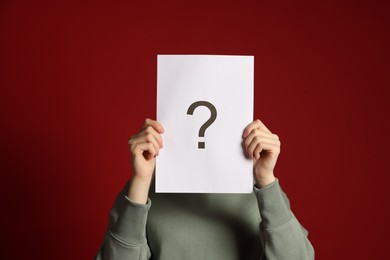 Photo of Anonymity. Woman hiding behind paper sheet with question mark on dark red background