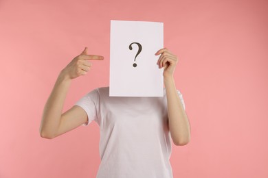 Photo of Anonymity. Woman hiding behind paper sheet with question mark on pink background