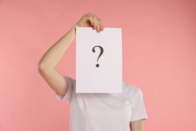 Photo of Anonymity. Woman hiding behind paper sheet with question mark on pink background