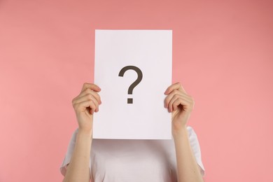 Photo of Anonymity. Woman hiding behind paper sheet with question mark on pink background