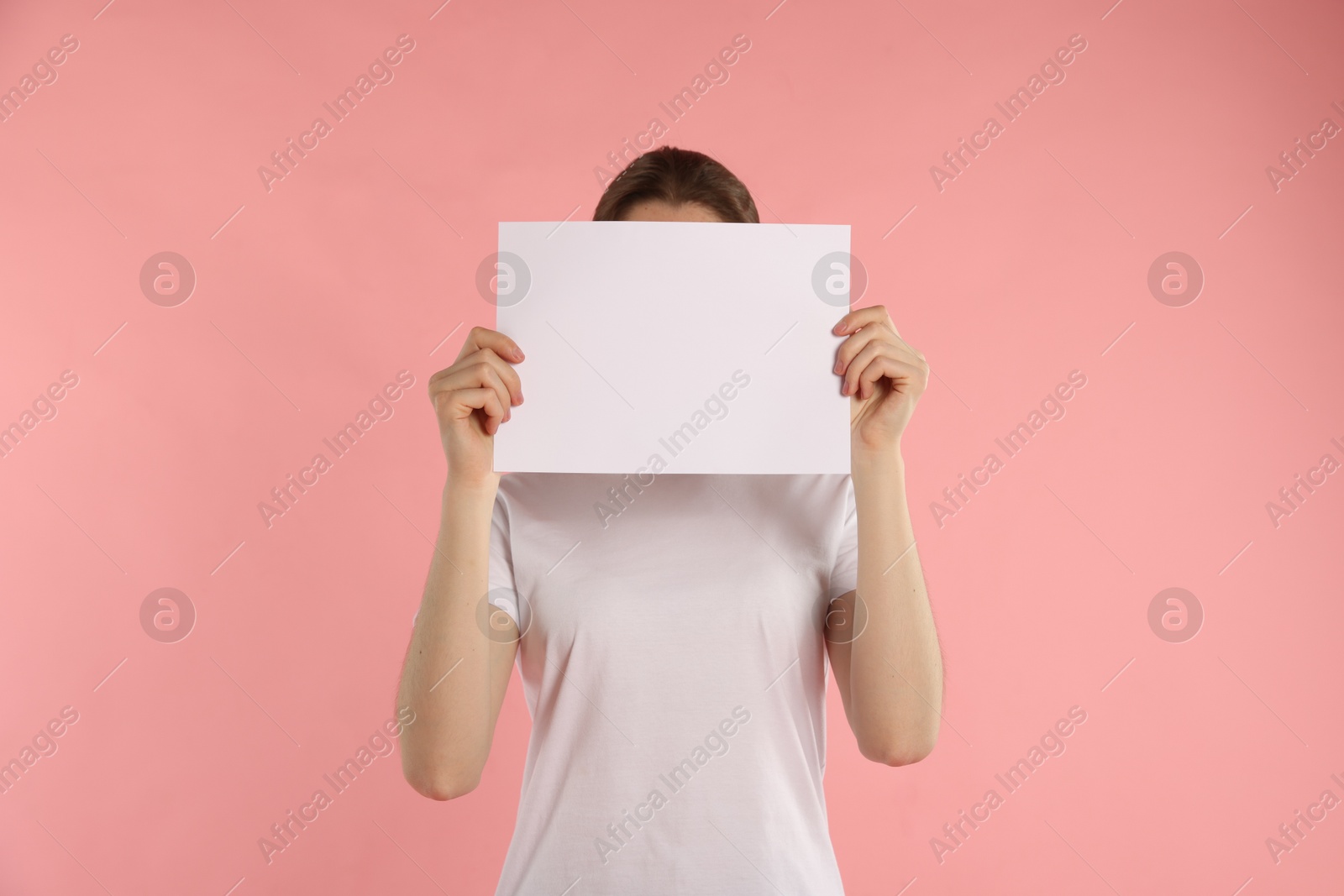 Anonymity. Woman hiding behind blank paper sheet on pink background Photo of Anonymity. Woman hiding behind blank paper sheet on pink background