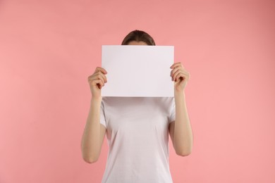 Photo of Anonymity. Woman hiding behind blank paper sheet on pink background
