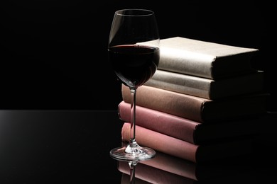 Photo of Red wine in glass and stack of books on mirror table against black background, closeup. Space for text