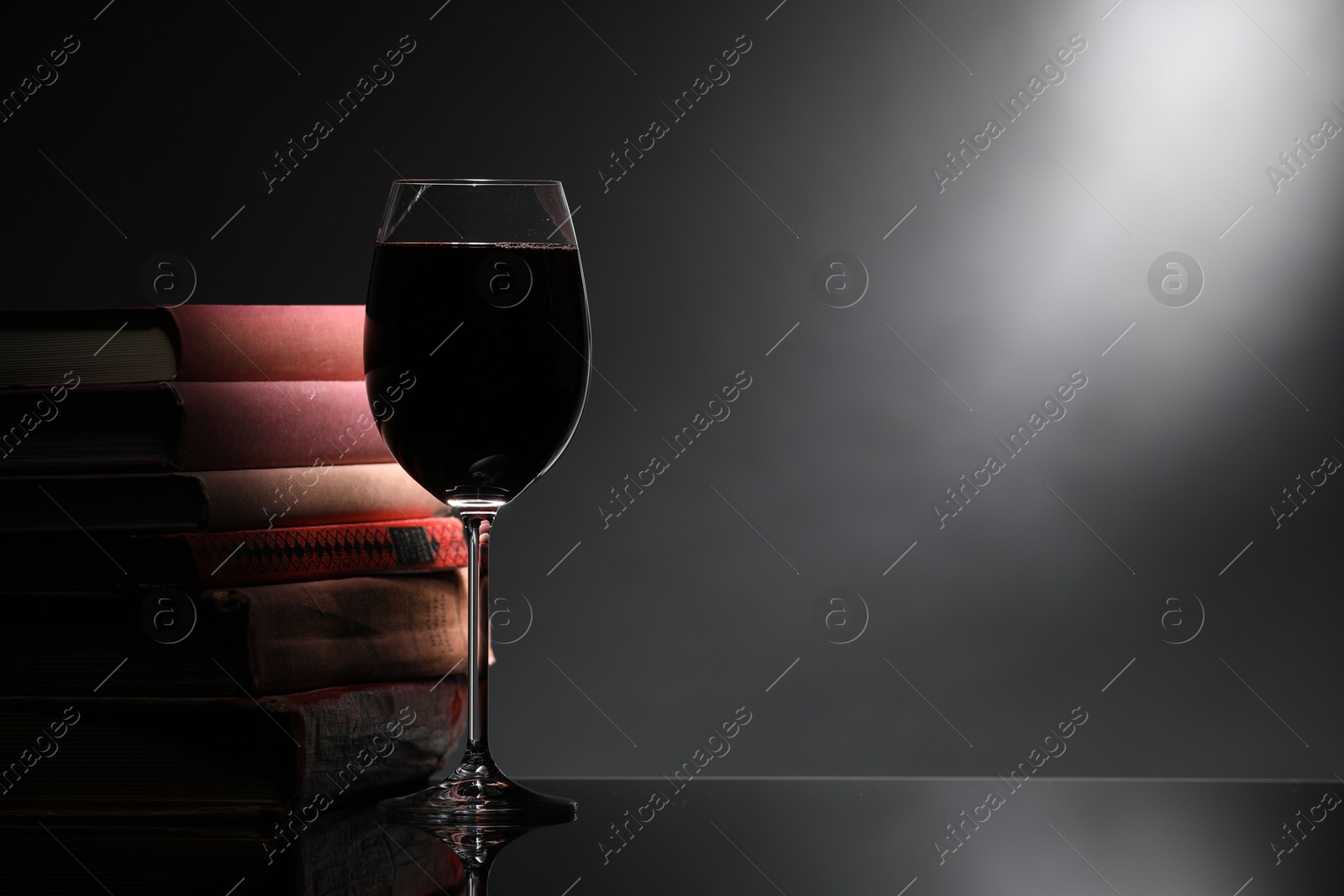 Tasty wine in glass and stack of books on mirror table against dark background. Space for text Photo of Tasty wine in glass and stack of books on mirror table against dark background. Space for text